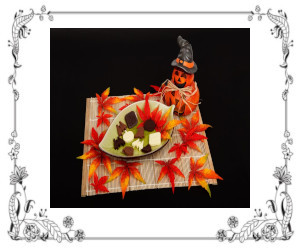 A jack o'lantern scarecrow sitting by a dish of Halloween candy dish with red maple leaves around it.
